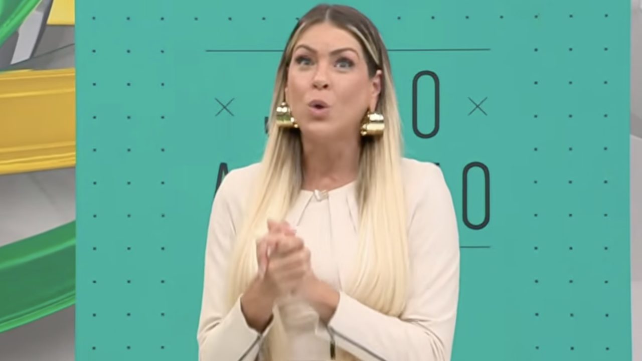 Renata Fan, de blusa clara e brincos dourados, com mãos entrelaçadas e expressão de surpresa no estúdio do Jogo Aberto diante de painel verde-turquesa