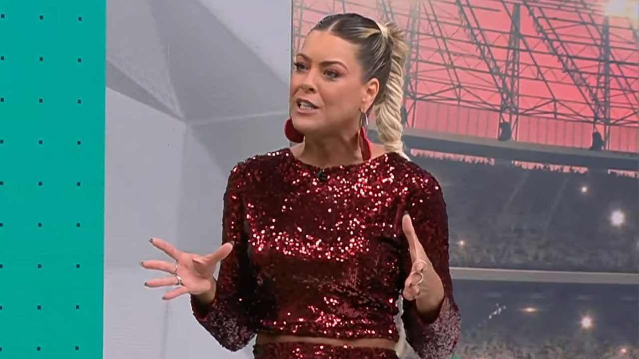 Renata Fan, apresentadora com cabelo loiro preso em trança alta, fala gesticulando com as mãos no estúdio do Jogo Aberto; ela veste blusa vermelha de mangas longas coberta de paetês e brincos grandes vermelhos, tendo ao fundo um painel com imagem de estádio e elementos gráficos em tons de verde e rosa.