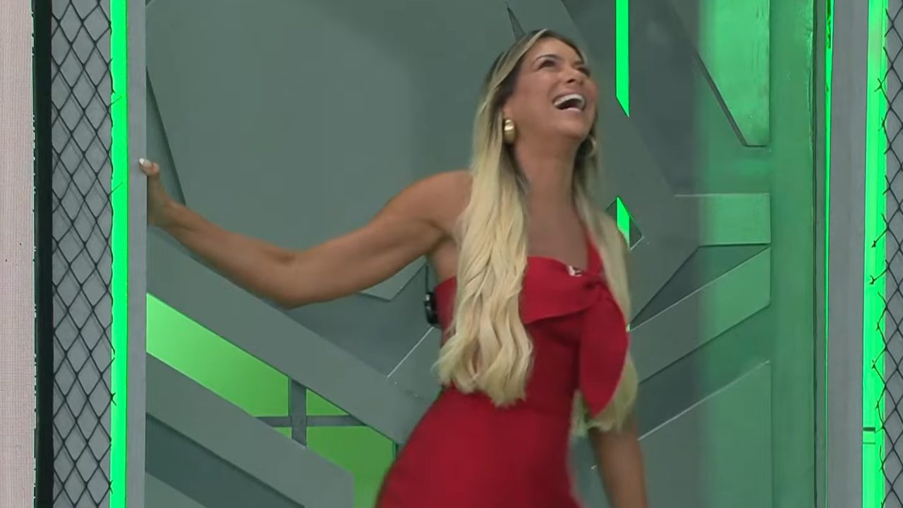 Renata Fan, de vestido vermelho, ri ao entrar no estúdio do Jogo Aberto, com iluminação verde e cenário moderno ao fundo.