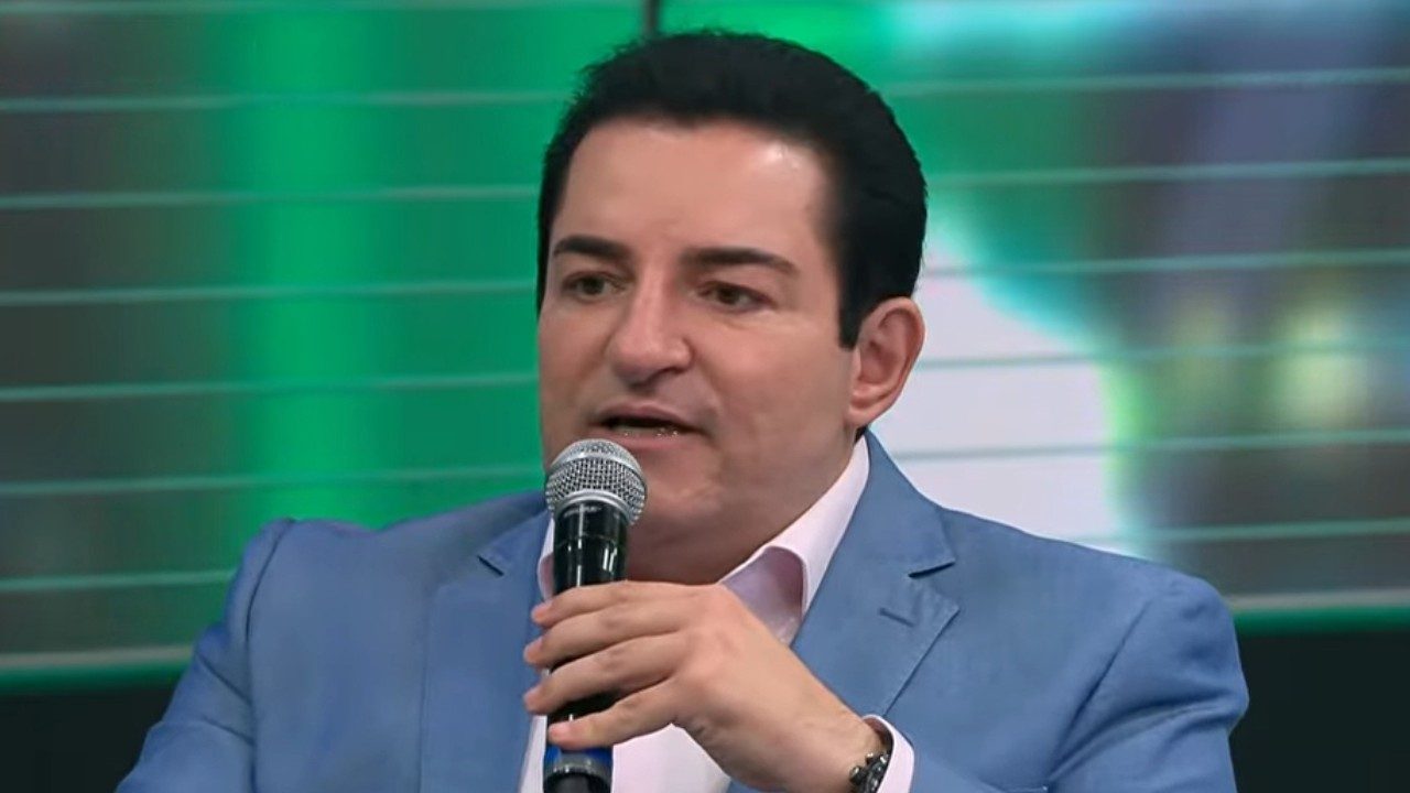 Renato Ambrósio fala ao microfone usando terno azul em estúdio de TV com fundo verde desfocado