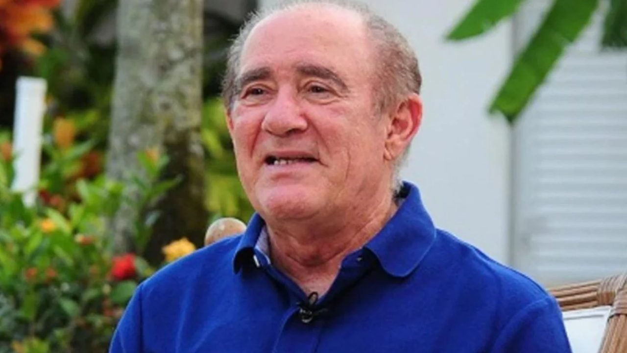 Homem de meia-idade veste camisa polo azul e sorri em ambiente externo, cercado por plantas e flores coloridas, transmitindo um clima leve e descontraído