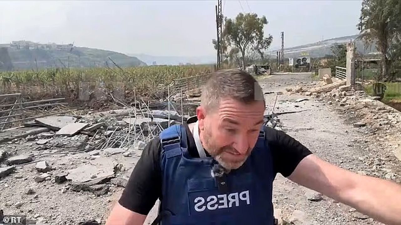 Um homem aparece em uma área externa com sinais de destruição, usando um colete à prova de balas com a palavra “PRESS”. Ele parece estar se abaixando ou se protegendo, com expressão tensa. Ao redor, há destroços, estrada danificada e vegetação ao fundo, sugerindo um cenário de conflito ou área recentemente atingida. A imagem transmite um momento de reportagem em situação de risco.