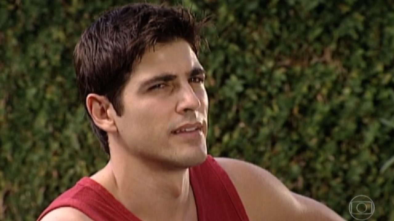 Homem jovem ao ar livre, vestindo camiseta regata vermelha, olha para o lado com expressão séria enquanto a luz do dia ilumina seu rosto. Ao fundo, há uma parede coberta por folhagem verde desfocada.