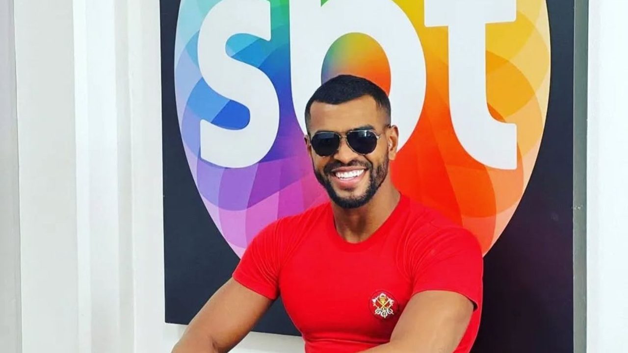 Homem sorri enquanto posa em frente ao logotipo colorido do SBT. Ele usa camiseta vermelha e óculos escuros, exibe barba bem aparada e está em um ambiente interno com fundo branco e o símbolo da emissora em destaque.