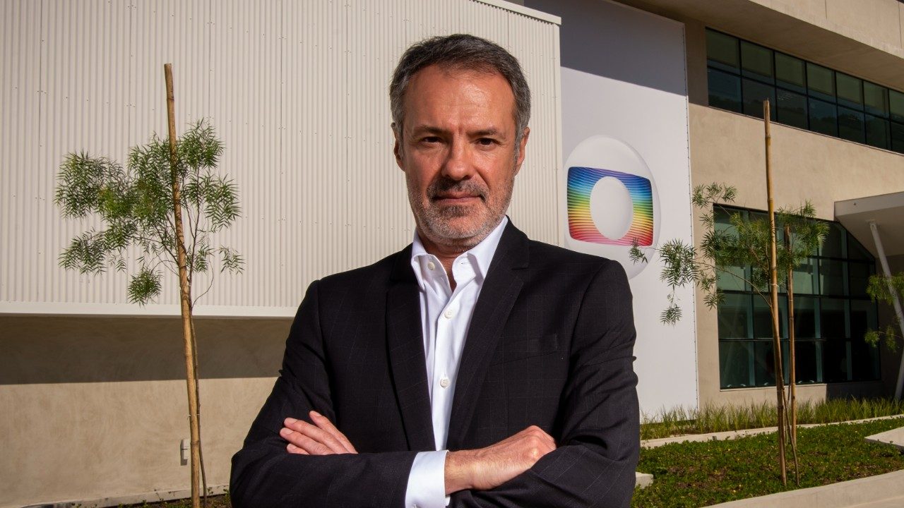 Ricardo Waddington de braços cruzados em frente a prédio dos Estúdios Globo com logotipo da emissora ao fundo
