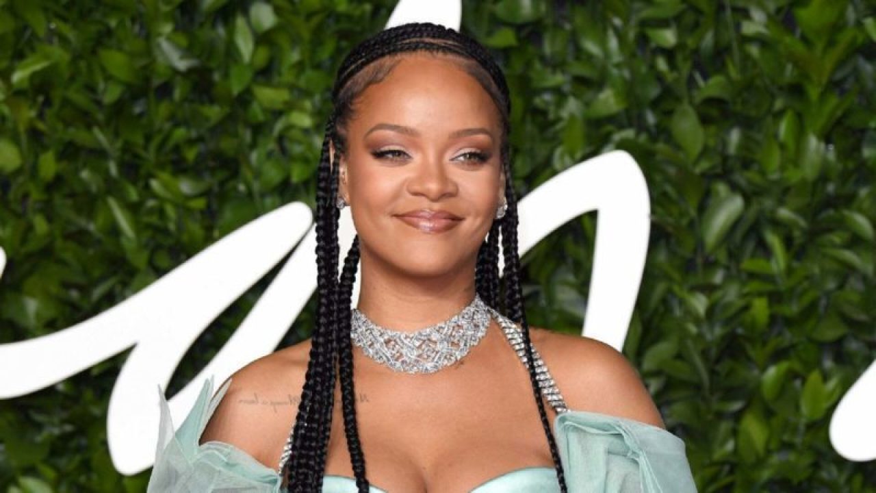 Rihanna usando vestido verde claro com colar brilhante em evento com fundo de folhagens e letreiro branco