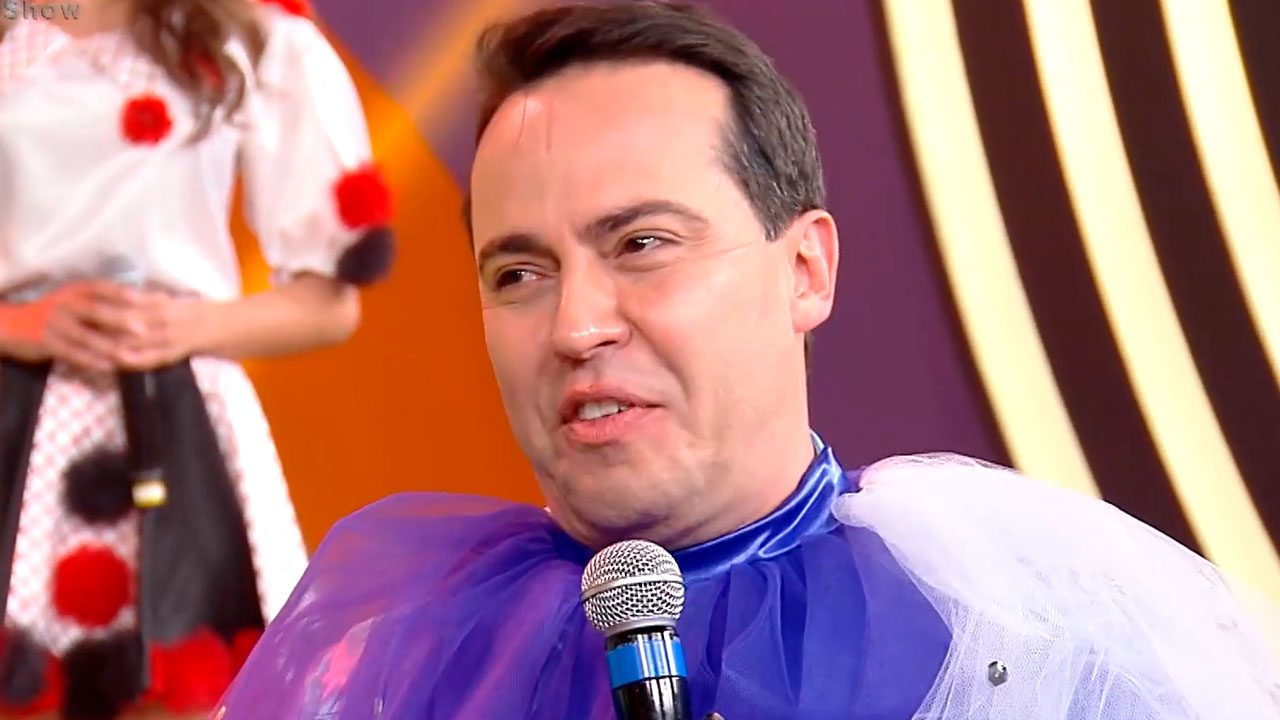 Rinaldi Faria fantasiado no palco do Domingo Show da Record em 2019 segurando microfone azul