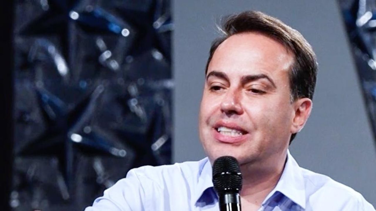 Homem de camisa social azul clara fala segurando um microfone em um ambiente interno, com painel decorado com estrelas ao fundo, sugerindo participação em um evento, palestra ou programa de TV