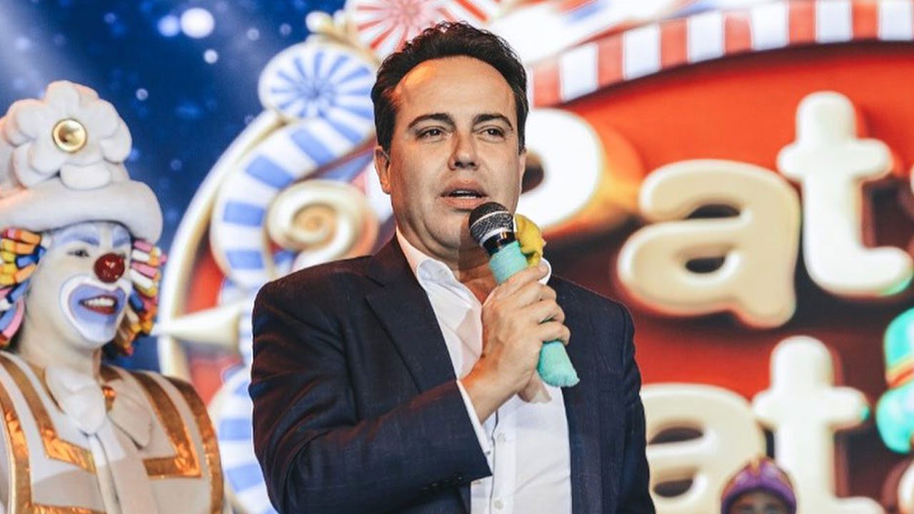 Rinaldi Faria, criador do Patati Patatá, falando ao microfone em um evento com fundo temático de doces e um palhaço