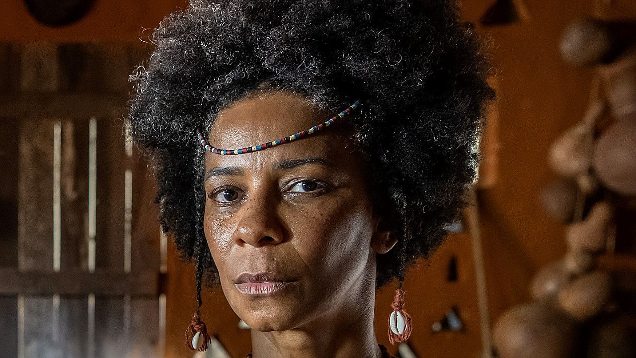 Rita Batista com adereço indígena e expressão séria em cena da série documental sobre ancestralidade