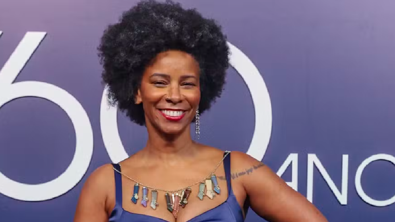 Rita Batista sorridente posa em frente a um painel roxo, vestindo um vestido azul e colar colorido, com cabelo em estilo afro e batom vermelho
