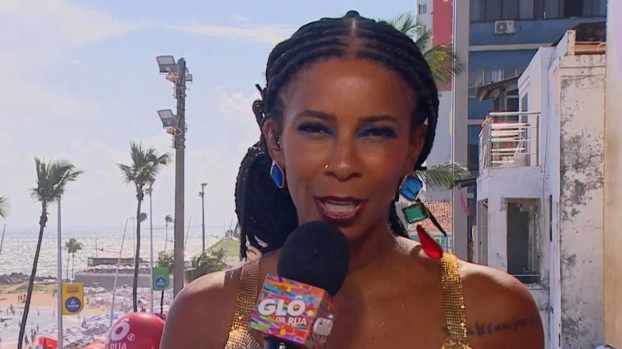 Mulher segura um microfone com o logotipo colorido do “GLO da Rua” e fala diante da câmera em um cenário ensolarado de praia, com o mar, coqueiros e prédios ao fundo. Ela usa vestido dourado brilhante, brincos grandes e coloridos, e maquiagem com sombra azul nos olhos.