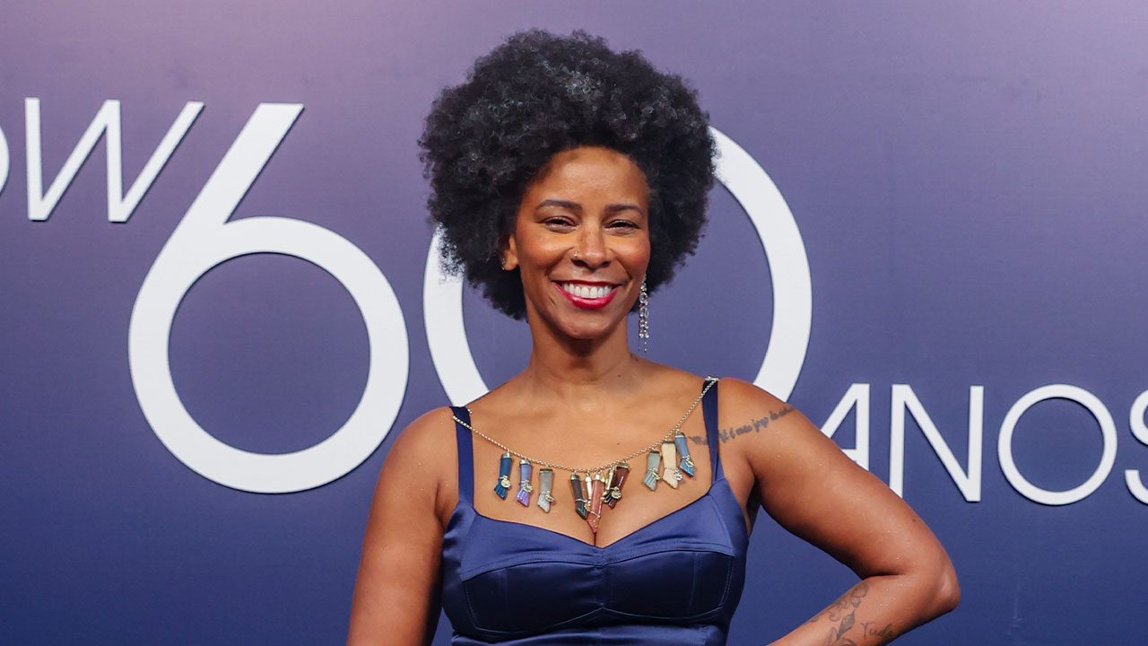 Rita Batista sorrindo em frente a painel da Globo durante evento de 60 anos da emissora