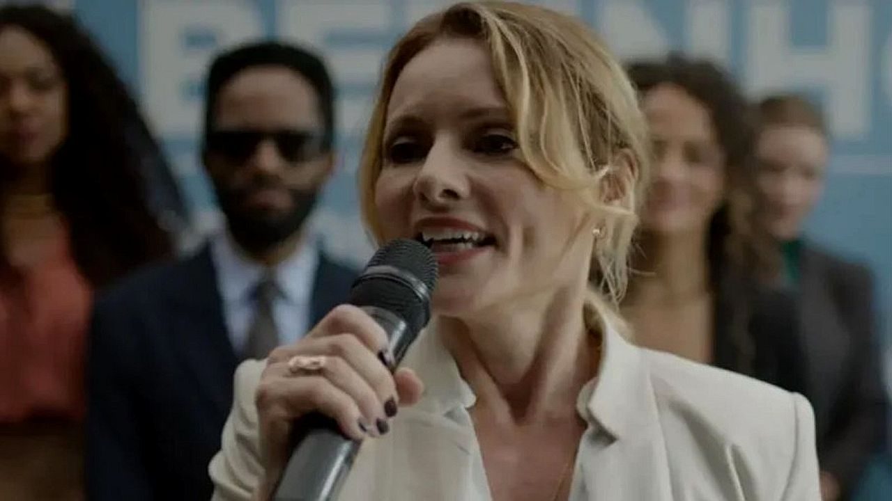 Mulher fala ao microfone em evento público, vestindo blazer claro e com expressão determinada. Ao fundo, outras pessoas aparecem desfocadas, sugerindo coletiva de imprensa, campanha ou anúncio oficial em ambiente corporativo.