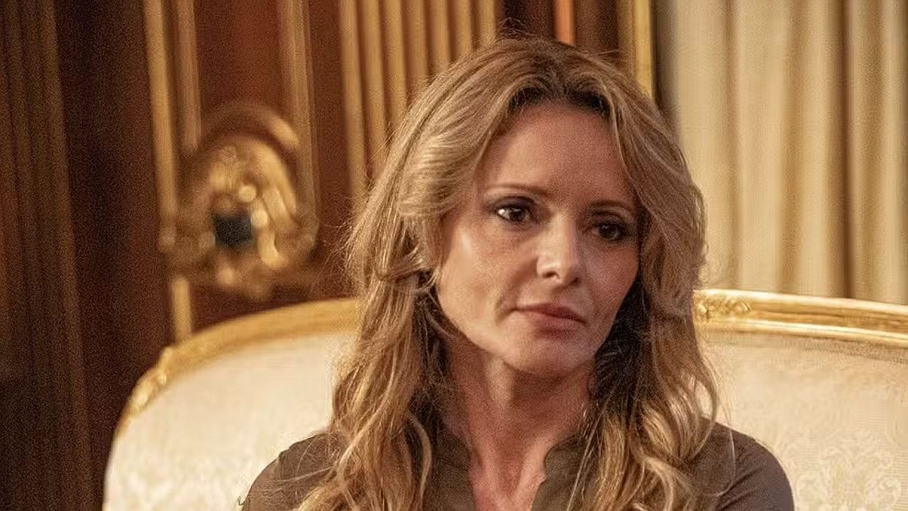 Rita Guedes com expressão séria sentada em poltrona elegante em cena de novela ambientada em mansão clássica