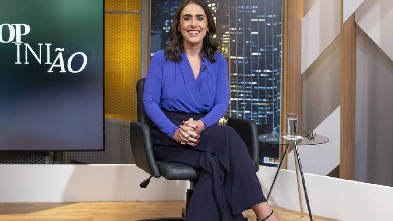 Mulher sentada em uma cadeira de estúdio televisivo sorri para a câmera. Ela veste blusa azul vibrante e calça escura, com sandálias de salto. Ao lado, há uma mesa pequena com um copo de água e óculos. Ao fundo, vê-se um painel com cidade iluminada à noite e a palavra “OPINIÃO”