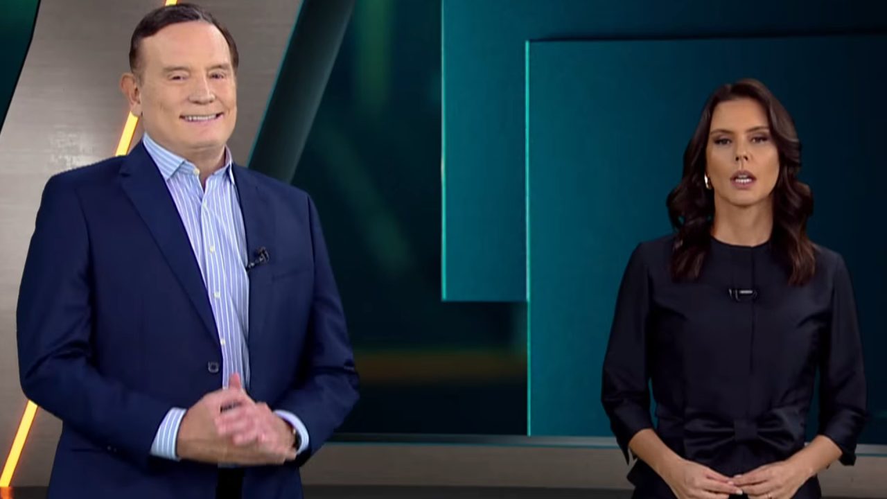 Roberto Cabrini e Camila Busnello, apresentadores do Domingo Espetacular, em pé no estúdio durante o programa, com um cenário moderno ao fundo.