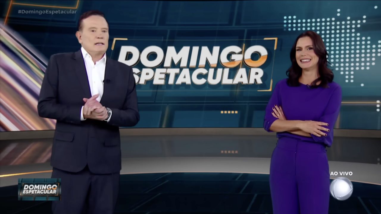 Roberto Cabrini, de terno, e Camila Busnello, de conjunto roxo, no estúdio do Domingo Espetacular diante do telão do programa