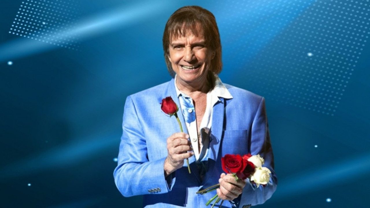 Cantor Roberto Carlos sorri segurando rosas vermelhas e brancas em imagem promocional associada ao especial da TV Globo