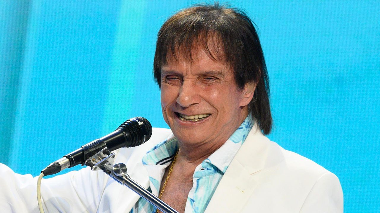 Roberto Carlos sorrindo em apresentação musical ao microfone com fundo azul durante especial de fim de ano na TV Globo