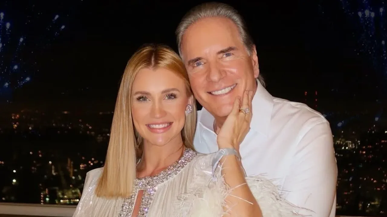 Na imagem, Roberto Justus e Ana Paula Siebert posam sorridentes e abraçados diante de uma vista noturna iluminada, com pontos de luz ao fundo que indicam a cidade e fogos de artifício no céu. Ana Paula usa um vestido branco com detalhes em brilho e plumas, enquanto Justus está com camisa social branca. Ambos exibem expressões de alegria e cumplicidade, reforçadas pela mão dela apoiada no rosto dele. O cenário e a produção visual sugerem um momento especial, possivelmente comemorativo.