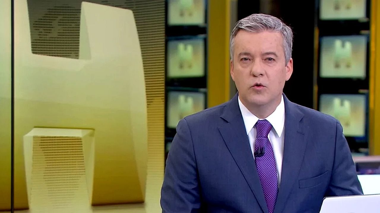 Âncora de telejornal apresenta notícia em estúdio com telões ao fundo e bancada de jornalismo