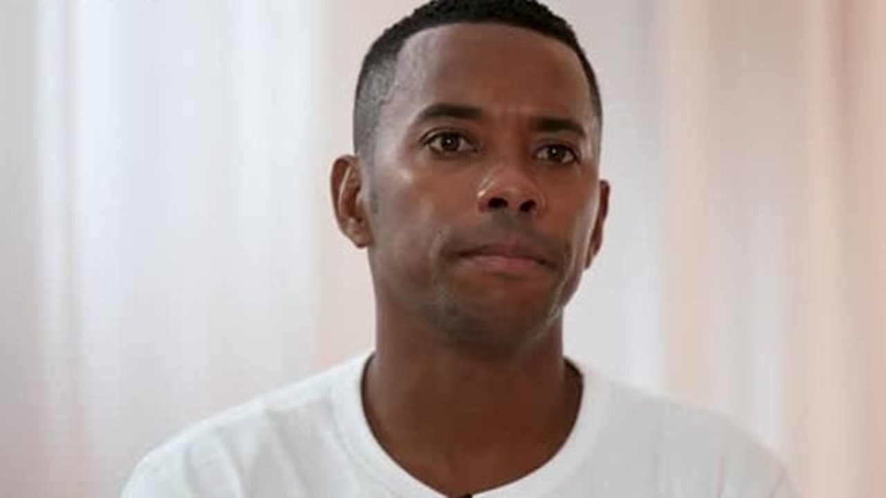 A imagem mostra um homem de expressão séria, vestindo uma camiseta branca, em frente a um fundo claro — possivelmente uma cortina. A iluminação é suave, e o clima da cena parece introspectivo ou reflexivo