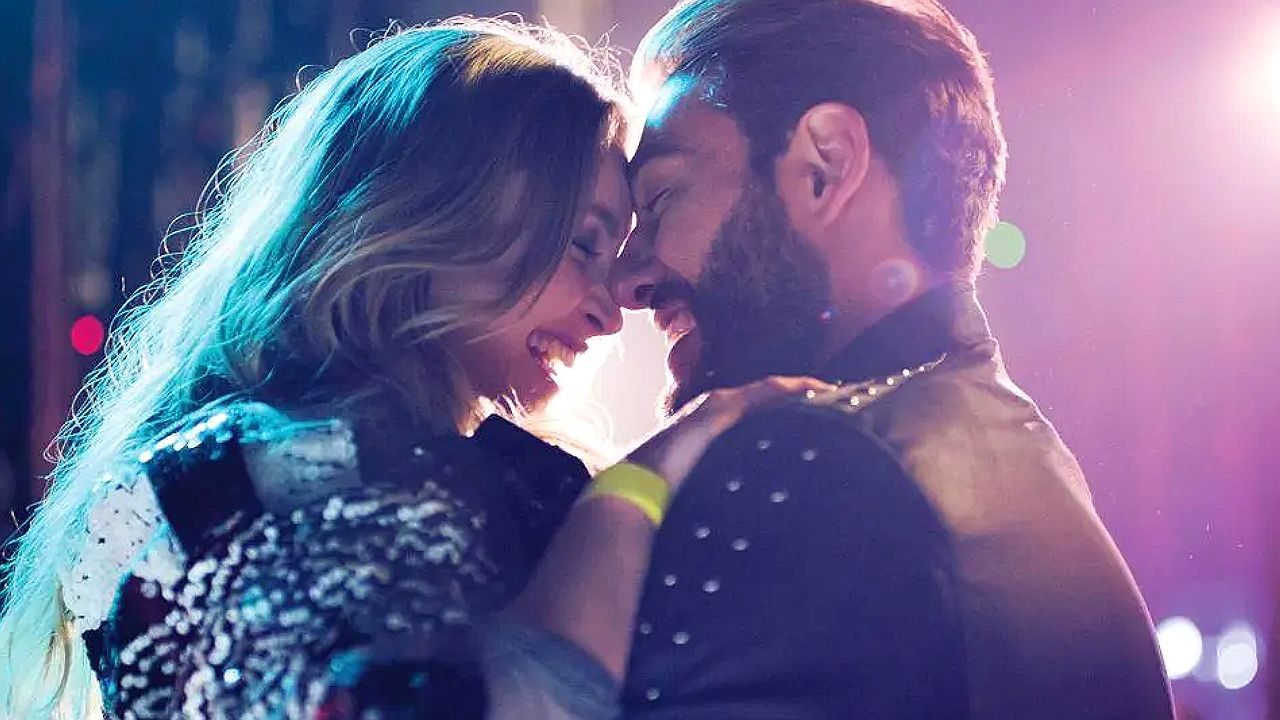 Casal se abraça e sorri durante momento romântico sob luzes coloridas em cena de filme ou série