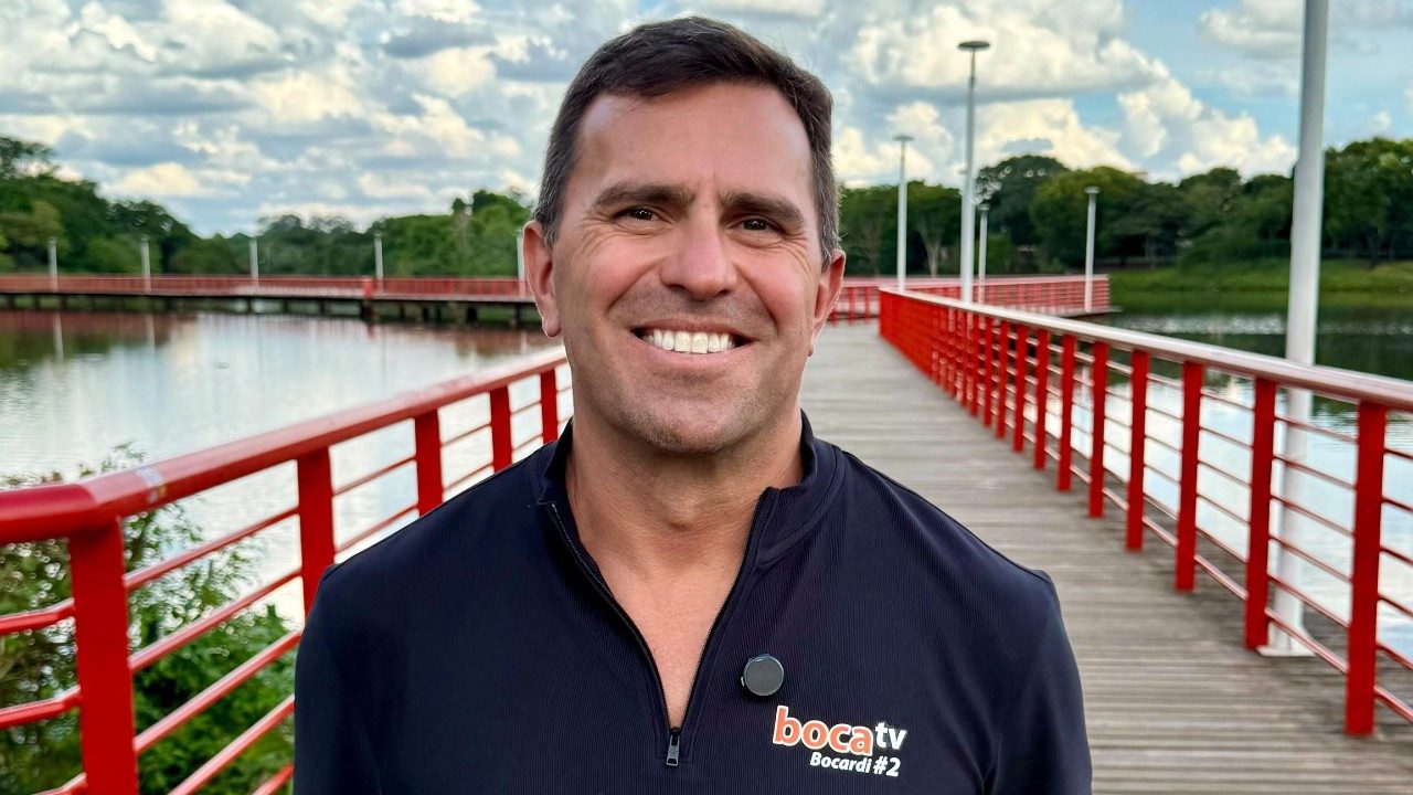 Rodrigo Bocardi sorri em passarela sobre lago usando blusa preta com logotipo da Boca TV ao ar livre