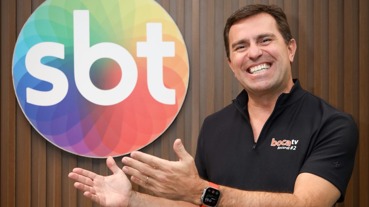Rodrigo Bocardi sorri e gesticula diante do logotipo do SBT em foto corporativa na sede da emissora
