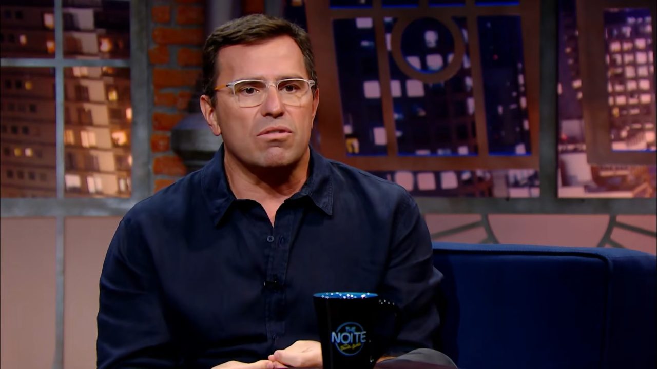 Rodrigo Bocardi sentado em sofá, em frente a uma mesa com caneca do The Noite, no cenário de talk show do SBT.