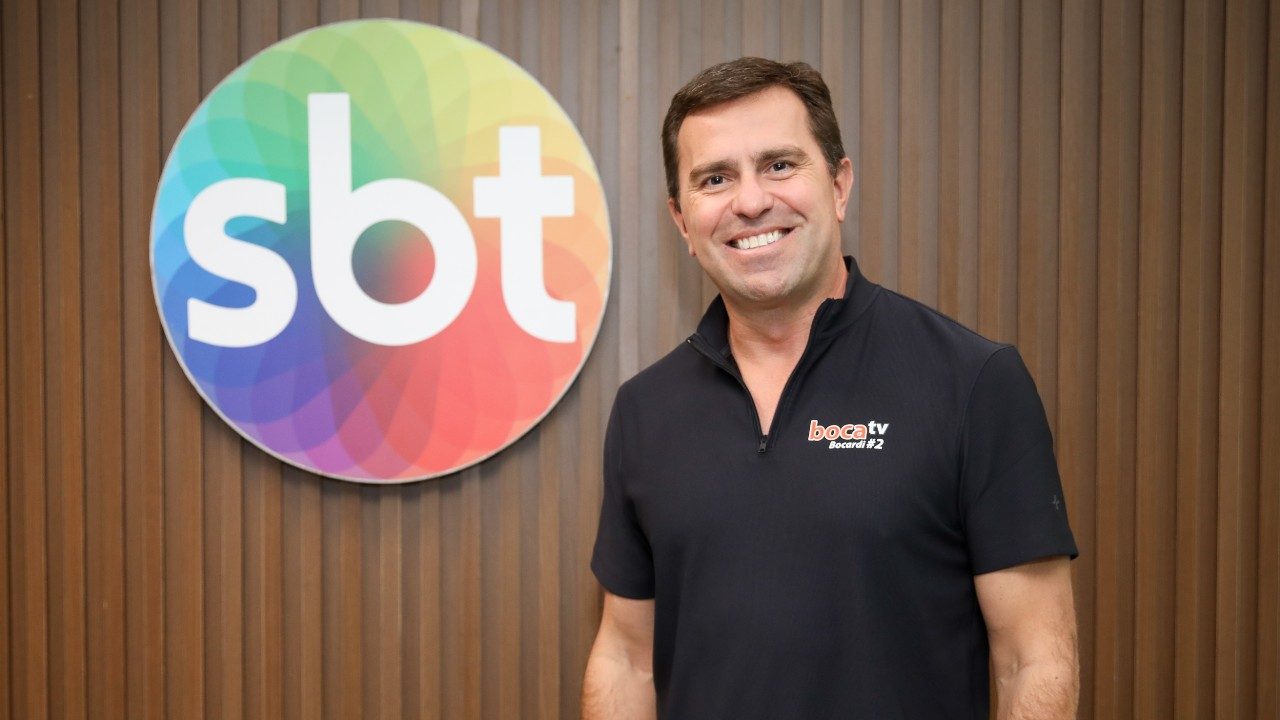 Rodrigo Bocardi sorri diante do logotipo do SBT, veste camisa preta com zíper e posa em frente a painel de madeira