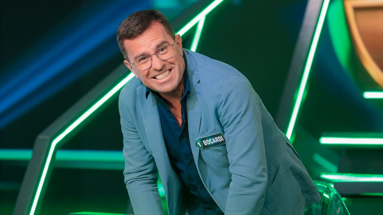 A imagem mostra Rodrigo Bocardi sorridente em um estúdio com luzes verdes e azuis. Ele usa óculos, blazer azul-claro e camisa azul-escura, com um crachá no peito que exibe o nome Bocardi. O ambiente, que é o cenário do Show do Milhão, tem aparência moderna, típico de um programa de TV de entretenimento ou competição