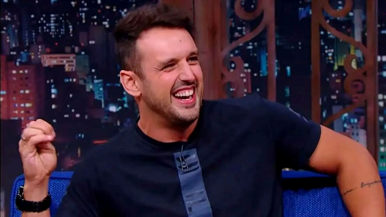 Rodrigo Capella ri durante entrevista em programa de auditório noturno com cenário urbano ao fundo