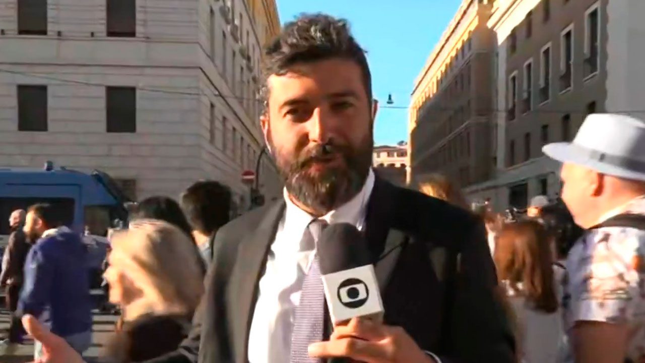 Rodrigo Carvalho, repórter da Globo, segurando microfone durante entrada ao vivo em rua movimentada no Vaticano