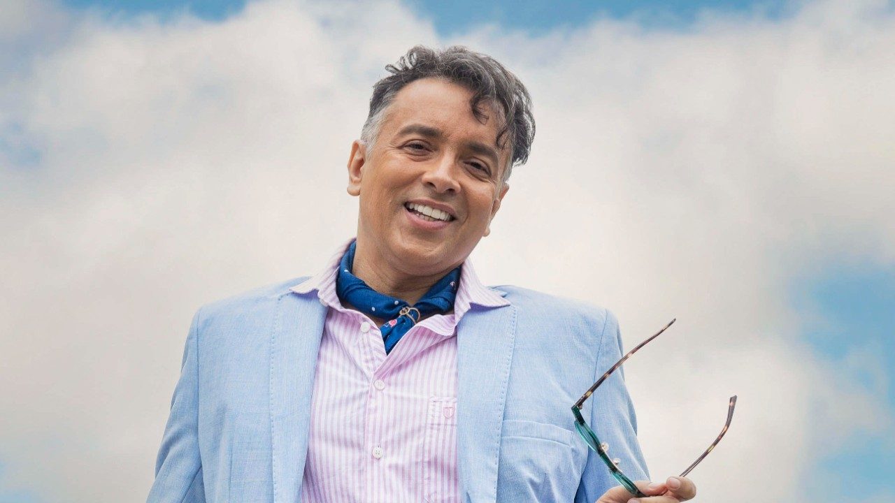 Ator Rodrigo Fagundes como Gigi na novela Volta por Cima, sorri usando blazer azul claro, camisa listrada rosa e lenço azul, sob um céu com nuvens