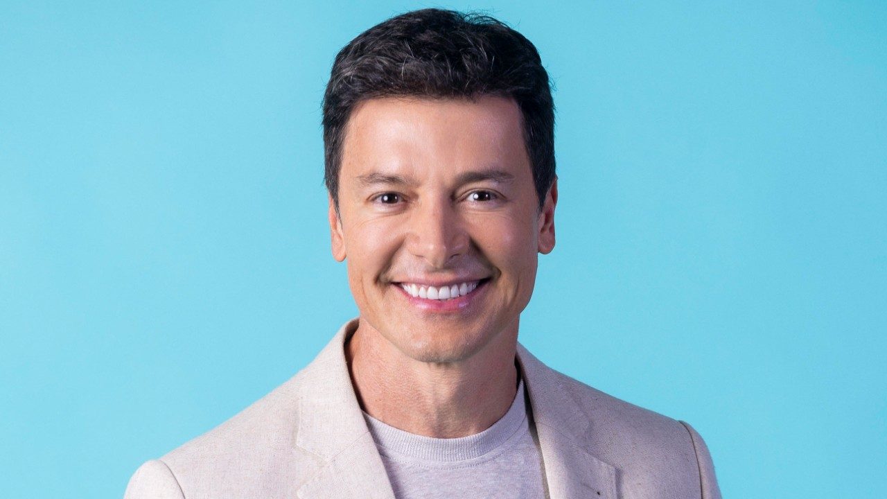 Rodrigo Faro sorri em retrato com fundo azul usando blazer claro e camiseta neutra em foto promocional