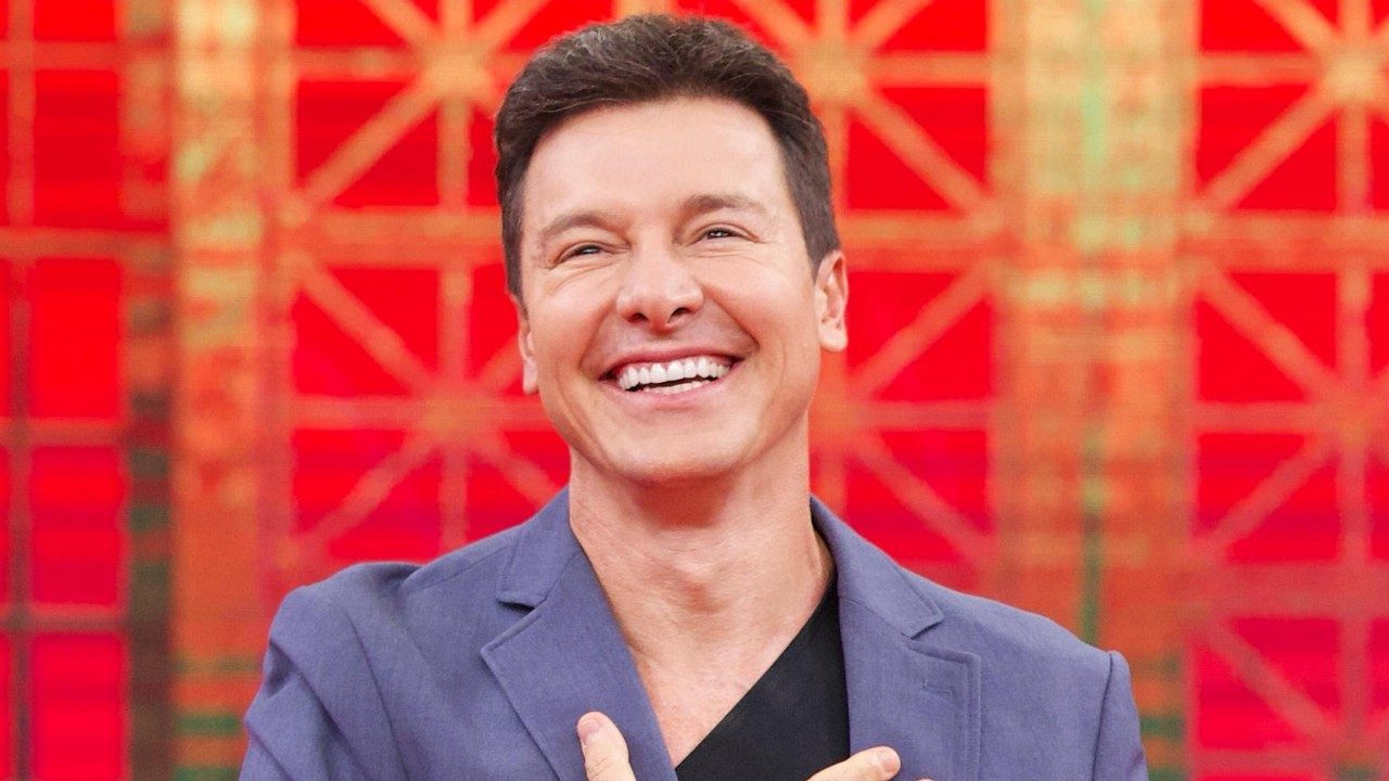 Rodrigo Faro sorrindo em estúdio com fundo vermelho geométrico durante gravação de programa de televisão