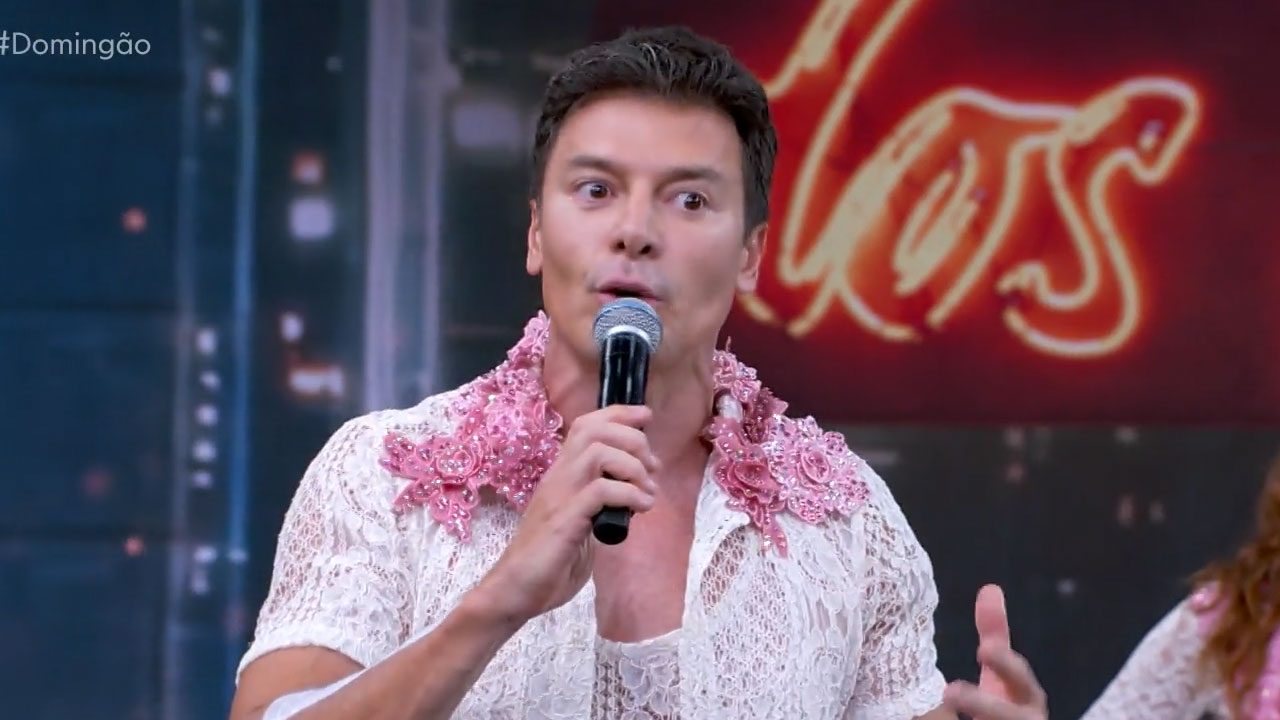 Rodrigo Faro com microfone na mão usando camisa rendada branca com flores rosas no ombro durante participação na Dança dos Famosos no Domingão