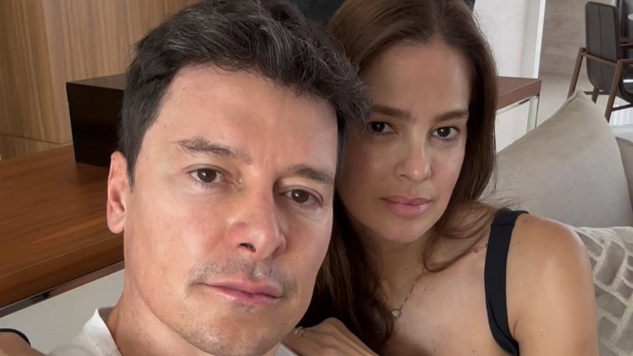 Homem e mulher sentados juntos em sofá de sala moderna posam para selfie com expressões sérias e olhar voltado para a câmera