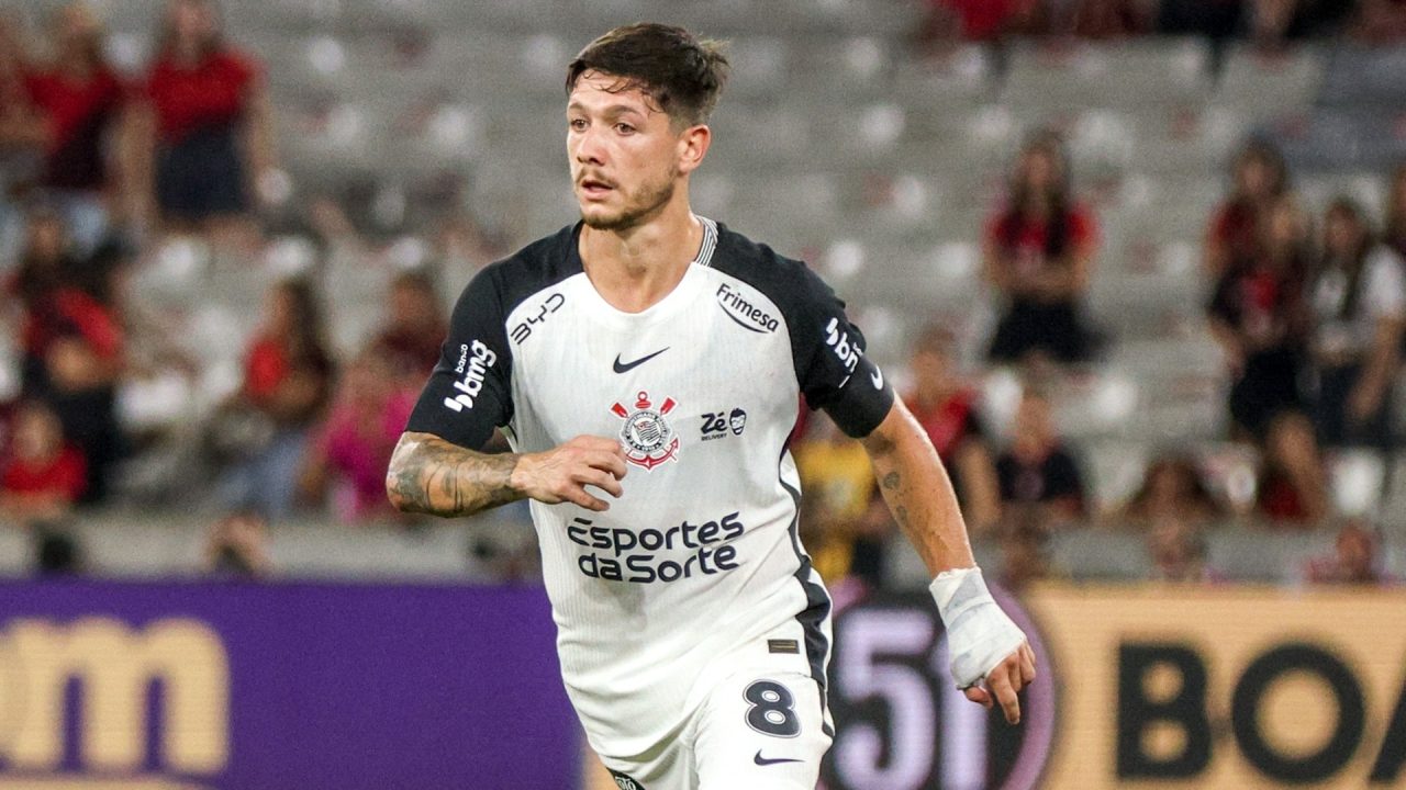 Rodrigo Garro, jogador de futebol com uniforme branco e preto do Corinthians, corre em campo durante partida, olhando para o lado; ele usa a camisa número 8, tem tatuagens no braço e a mão direita enfaixada, com arquibancadas desfocadas ao fundo.