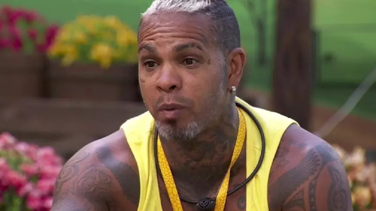 Rodriguinho em um momento de conversa ao ar livre, usando regata amarela e colar preto, com expressão séria e flores coloridas ao fundo