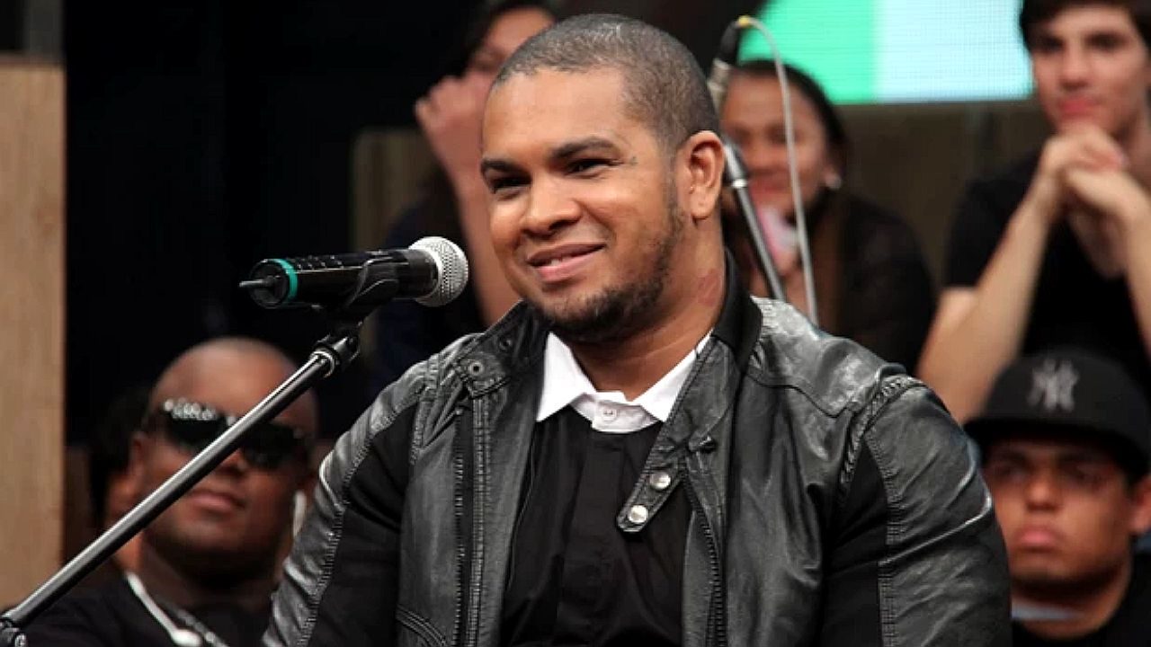 Homem sorri enquanto fala ao microfone durante apresentação ou entrevista em programa de televisão, com plateia e músicos ao fundo.