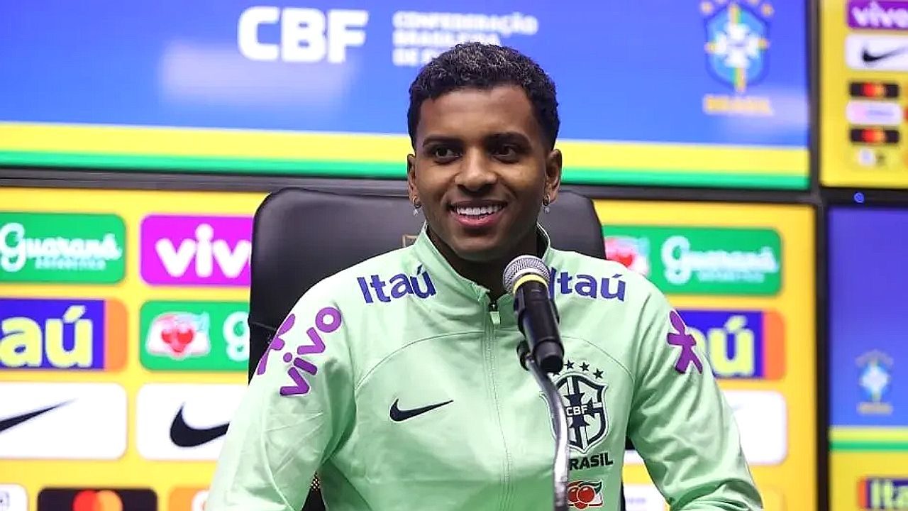 Jogador da seleção brasileira sorri durante entrevista coletiva com painel de patrocinadores ao fundo