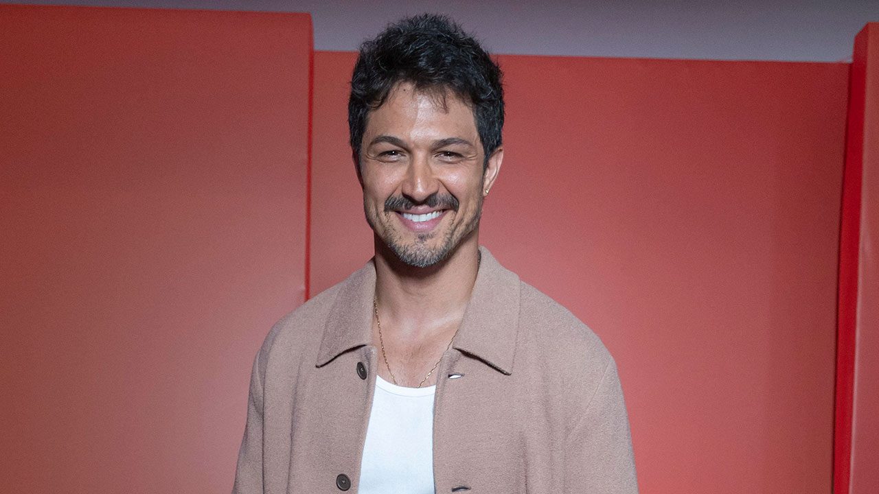 Romulo Estrela sorrindo com casaco bege diante de fundo vermelho em evento da Globo