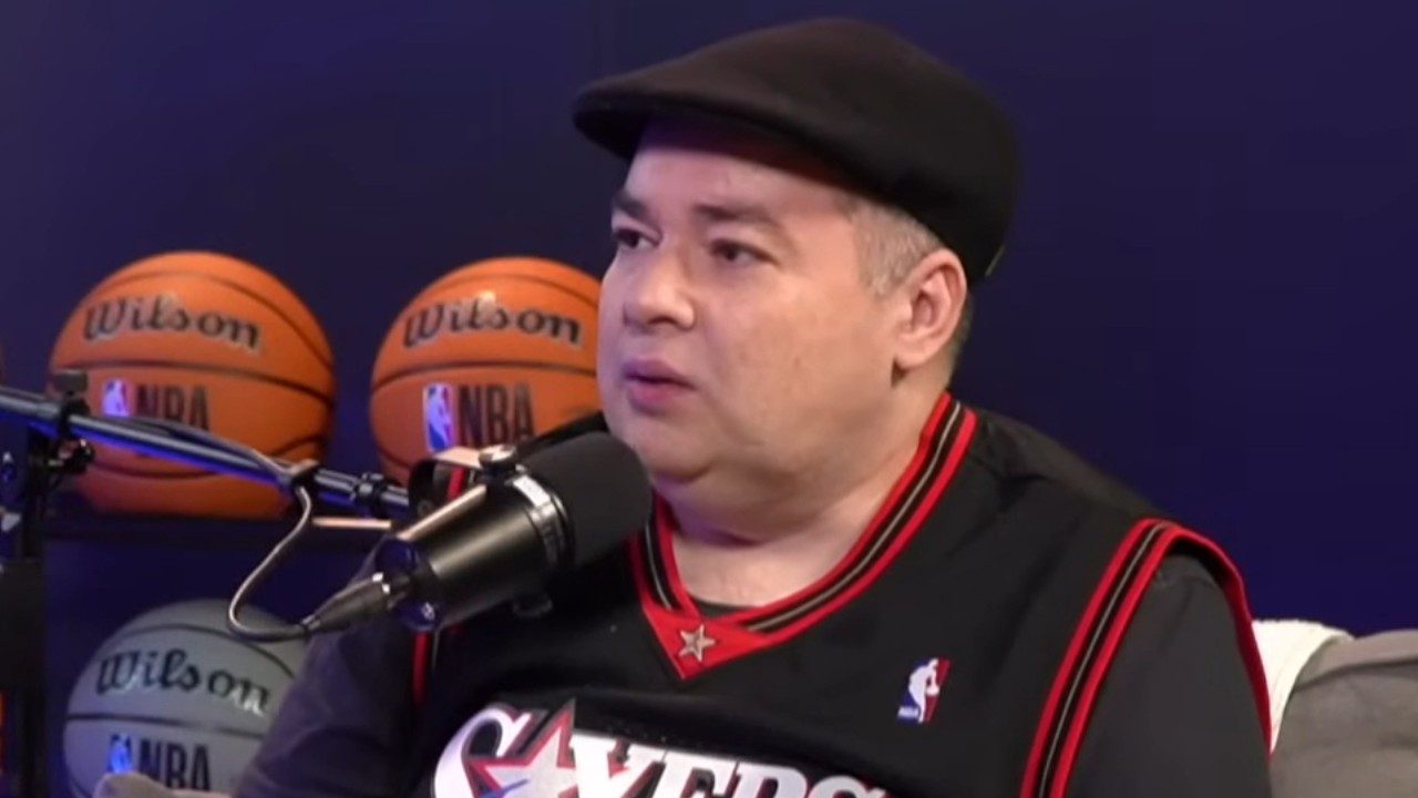 Rômulo Mendonça de boné preto e camisa da NBA fala ao microfone em estúdio com bolas de basquete ao fundo