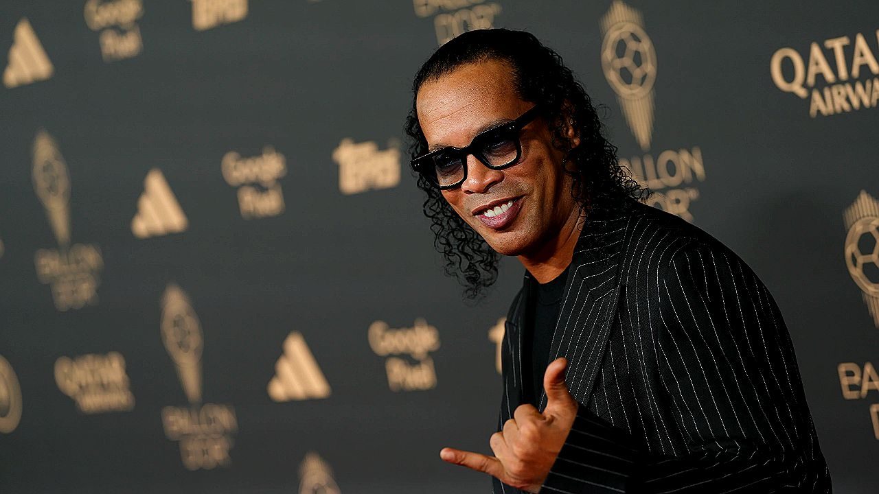 Ronaldinho Gaúcho posa sorrindo com gesto de mão em evento da Bola de Ouro com logos ao fundo