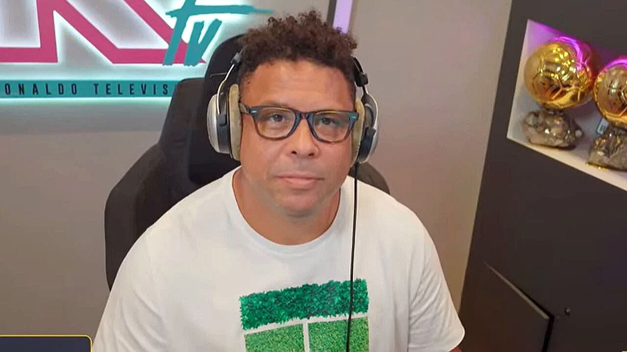 Homem de óculos e fones de ouvido participa de transmissão esportiva em estúdio com troféus ao fundo