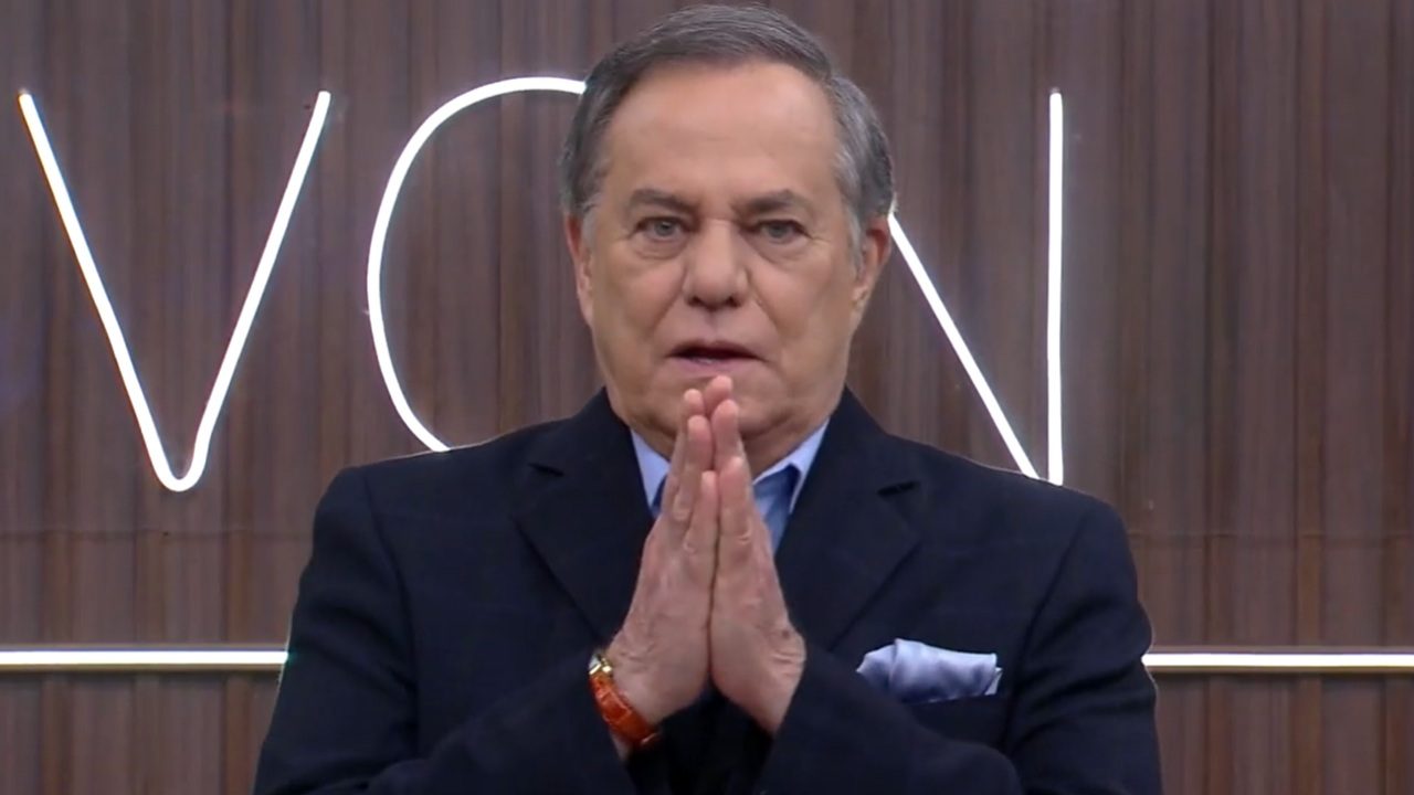 A imagem mostra um homem de cabelo grisalho, vestindo um terno escuro e camisa azul clara. Ele está com as mãos unidas em frente ao rosto, em um gesto que pode indicar concentração ou ênfase no que está dizendo. Ao fundo, há um painel de madeira com letras iluminadas, sugerindo que ele está em um estúdio de televisão