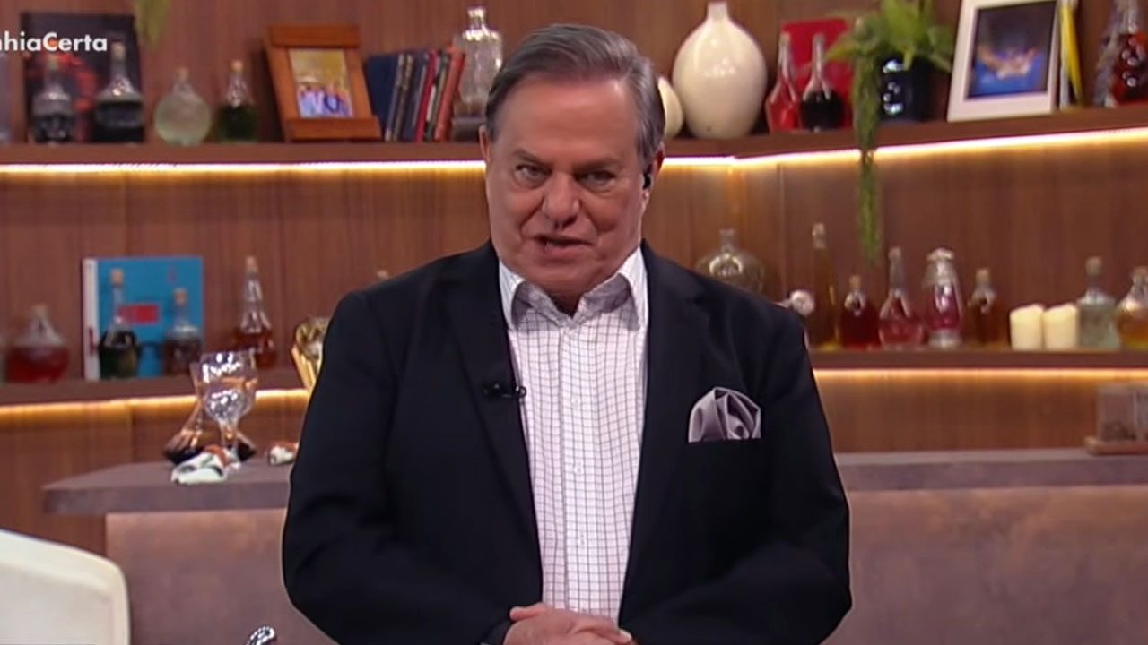 Ronnie Von em pé no estúdio do programa Companhia Certa com estante ao fundo exibindo garrafas objetos decorativos e iluminação cênica